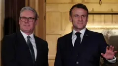 Thủ tướng Anh Keir Starmer và Tổng thống Pháp Emmanuel Macron, cả hai đều mặc vest và đeo cà vạt, đứng cạnh nhau tạo dáng trước ống kính của các nhiếp ảnh gia.