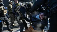 Agentes de ICE tratan de reducir a una persona que grita con una rodilla en el suelo.