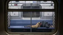 Homem sem-teto dormindo deitado no assento de um vagão parado na estação Jamaica Center do metrô de Nova York (EUA), uma das linhas mais usadas pelos sem-teto para refúgio na cidade, em Queens, Nova York, no dia 7 de abril de 2025