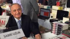 Sabri Uzun kitap fuarında, masada oturuyor, kameraya gülümsüyor