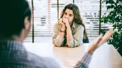Uma mulher em um escritório está sentada em frente a um homem, que aparece de costas, desfocado ao fundo; ela tem expressão de irritação, e ele gesticula