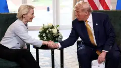 Von der Leyen, de jaqueta branca curta e calça preta, estende a mão diante de uma mesa lateral com um arranjo de flores brancas para cumprimentar Trump, que usa terno azul, gravata dourada e segura papéis na outra mão.