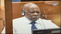 Félicien Kabuga imbere y'urukiko i La Haye mu Buholandi yambaye ishati y'umweru na karuvati y'ubururu n'umweru afite utwuma dufasha kumva mu matwi