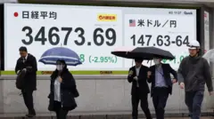 Pessoas caminham na rua em frente a tela com índice Nikkei