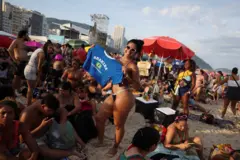 Mulher de biquíni segura camiseta com o nome Shakira e uma bandeira do Brasil. Está na praia, de dia.