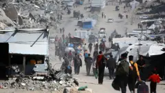 فلسطينيون يحملون أمتعتهم أثناء فرارهم من مدينة غزة، ويحيط بهم خيام وركام مبانٍ.