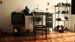 Ventilador apoiado em 'esqueleto' de cadeira; ao lado de armário de cozinha, há uma sapateira