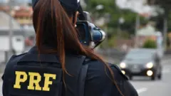 Agente da PRF, identificada por colete e boné, usa equipamento de fiscalização em estrada