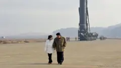 Com os longos cabelos presos, calças pretas e uma jaqueta acolchoada branca, Kim Ju Ae caminha de mãos dadas com Kim Jong Un, que veste uma jaqueta marrom e calças pretas. Eles se olham. Imponente acima deles, com vários homens ao lado e colinas ao fundo, um míssil balístico intercontinental (ICBM) aponta para cima, sobre um transportador de esteiras.