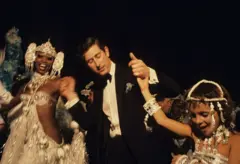 Rei Charles usando smoking ao lado de duas passistas de samba 