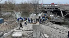 People fleeing Irpin cross the damaged river bridge, which can be seen with the concrete collapsed and girders hanging down into the water
