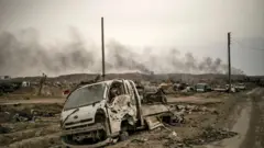 الدخان يتصاعد من خلف المركبات المدمرة والمباني المتضررة في قرية الباغوز بمحافظة دير الزور شرقي سوريا