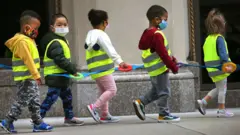 Des enfants d'une classe de maternelle portent des gilets et des masques et s'accrochent à une sangle lors d'une promenade en plein air à Boston.