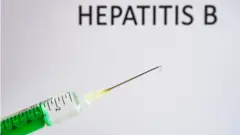 Di foto illustration show disposable syringe with hypodermic needle, dem write HEPATITIS B on a white board behind.