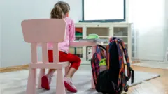 Em casa, menina sentada diante de mesa e TV, com mochila ao lado