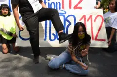 Um manifestante coloca o pé no ombro de uma jovem manifestante que está ajoelhada no chão, amordaçada com uma corda amarrada nos braços e no peito. Uma faixa pintada atrás deles exibe a Lei 112, a seção da lei tailandesa que criminaliza insultos ao monarca.