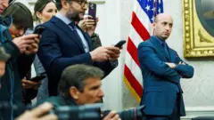 Stephen Miller stands in the Oval Office next to the press who are pointing cameras at Trump who is off camera. He is standing with his arms folded