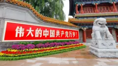 Um letreiro vermelho com caracteres chineses brancos está pendurado em um muro de pedra, com um colorido canteiro de flores abaixo; um leão de pedra branca está em cima de um pedestal ao lado, enquanto um edifício ornamentado aparece ao fundo.