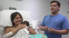 अस्पताल के बिस्तर पर लेटी डायना अपने बेटे जुआन डेविड को अपनी छाती से लगाए हुए मुस्कुरा रही हैं. उनके पति, गेरसन, हल्के नीले रंग की टी-शर्ट पहने उनके पास खड़े हैं.