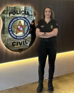 A delegada Amanda Souza o lado de um logotipo da Polícia Civil. Ela veste roupas pretas da polícia e calça bota e está com os braços cruzados, sorrindo de pé para a câmera
