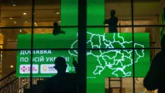 Великий зелений світлодіодний екран у формі аптечного хреста з картою України та написом «Доставка по всій Україні». Перед екраном видно силуети людей, які, ймовірно, встановлюють конструкцію.