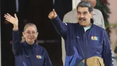 Cilia Flores saluda al parecer a una multitud con el brazo extendido. A su lado, Nicolás Maduro señala a un punto fuera del plano. Ambos sonríen.