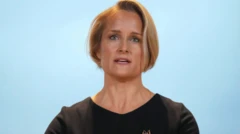 Metreweli kürsünün arkasında konuşma yapıyor. Üzerinde siyah bir elbise ve bir broş var. 