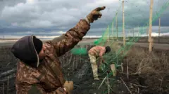Двое украинских военных натягивают антидроновую сетку на столбах вдоль дороги 