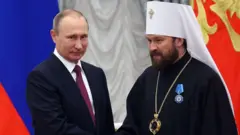 Путін (ліворуч) в темному костюмі та краватці та Іларіон (праворуч) – в чорному релігійному вбранні з білим головним убором, із великим хрестом на ланцюгу на грудях і нагородою на одязі – стоять поруч у приміщенні перед великими прапорами РФ і декоративним інтер’єром.
