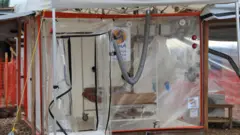 Uma estrutura transparente de plástico em formato de cubo, com uma porta de zíper aberta. Há uma cama vazia no interior e nenhum paciente
