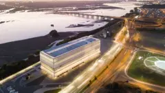 Edificio moderno junto a una carretera iluminada al atardecer, con un río amplio y puentes al fondo.