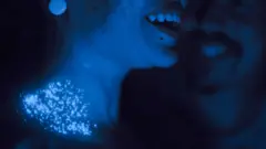 Cena de Emergência Radioativa mostra personagem em iluminação escura, com o rosto parcialmente visível e a boca entreaberta, enquanto pequenas partículas brilhantes — césio-137 — se espalham e reluzem na região de seu pescoço.