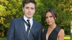 Brooklyn Beckham standing with Victoria Beckham in an outside setting with trees and a lawn in the background. They are in evening dress 