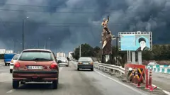 Veículos trafegam por uma rodovia, passando por uma estátua em memória aos mortos na guerra e um outdoor retratando o falecido líder supremo do Irã, o aiatolá Ali Khamenei, morto em um ataque aéreo em 28 de fevereiro, com colunas de fumaça preta subindo, em Teerã, em 8 de março.