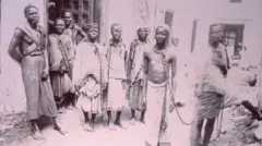 Fotografia do século 19 mostra pessoas negras escravizadas em correntes em um mercado na ilha africana de Zanzibar, aparentemente olhando para a câmera