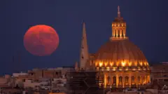 Uma Lua cheia, grande e avermelhada surge logo acima de edifícios no horizonte em Malta, com uma torre de catedral e uma cúpula de basílica ao lado