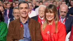 Kırmızı rahat giysi giymiş bir kadınla, yine rahat bir blazer ceket ve mavi gömlek giymiş bir adam yan yana oturuyor. Arkalarındaki koltuklar da dolu