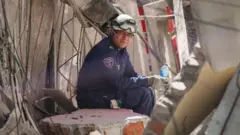 A rescue worker in a blue outfit and white helmet sits amid rubble