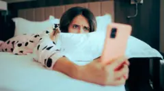 Mulher deitada na cama, olhando apreensiva para o telefone celular