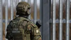 Soldado armado da Polônia, com identificação nas costas de que se trata de um guarda de fronteira, vigia uma barreira metálica