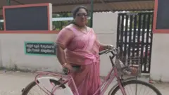Jayachithra, vestindo um sári rosa e segurando uma bicicleta feminina rosa, em frente a uma casa