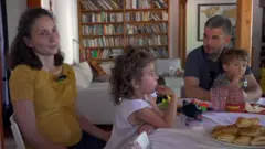 Egy férfi és egy nő ül egy asztalnál, mindegyikük ölében egy-egy kisgyerekkel. Az asztalon péksütemények és játékok. A háttérben egy könyvespolc látható, és egy kanapé rajta egy gitárral.
