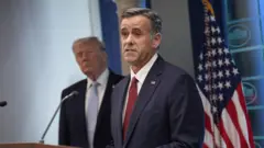 El director de la CIA, John Ratcliffe, hablando frente a un micrófono en la Casa Blanca, con Donald Trump detrás suyo. A su lado hay una bandera de Estados Unidos.