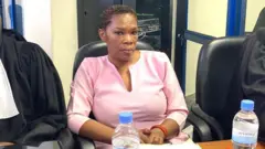 Béatrice Munyenyezi yicaye ku ntebe yegamirwa y'umukara, yambaye ikanzu y'ibara ry'iroza, akikiye ikarine (carnet) ku bibero ayifasheho n'ibiganza byombi. Imbere ye hari amacupa abiri y'amazi ya kompanyi Inyange. Munyenyezi akikijwe n'abanyamategeko babiri bambaye imyenda y'umukara, mu maso yabo ntihaboneka kuri iyi foto yo mu mubiko.