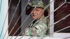 Atrás de uma grade e apoiado no parapeito de uma janela, Edith olha para a câmera enquanto exibe um uniforme e um chapéu militar.