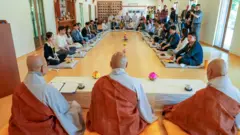 Grande grupo de participantes do evento sentados sobre almofadas em um amplo semicírculo no interior da sala de um templo, em frente a três monges