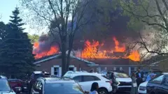 Abantu bateraniye ahantu, mu gihe umwotsi uzamuka hamwe n'umuriro, nyuma y'ibyabaye, aho umugabo yinjije imodoka ye mu muryango w'urusengero i Michigan akarashisha imbunda ndetse agatwika urusengero, mu mujyi wa Grand Blanc, muri leta ya Michigan, muri Amerika, ku itariki ya 28 Nzeri (9) mu mwaka wa 2025. 