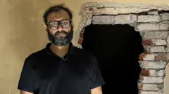 Mir Bin Ahmad Quasem stands in a detention centre looking at the camera, he wears glasses, a dark polo shirt and light trousers and has a beard. He is stood in front of a hole in the wall exposing inner brickwork with a pile of rubble just in frame to his right.
