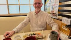 Bjørn-Eirik Olsen, sentado à mesa com um prato de sushi à sua frente. Ele veste uma camisa branca e sorri para a câmera.