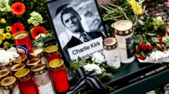 Retrato de Charlie Kirk em preto e branco rodeado de velas e flores.